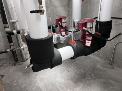 Insulated white-jacketed pipes with red circulator pumps and black insulated elbows installed by Metro Insulation White insulated pipes, red circulator pumps and black insulated pipe elbows in a mechanical room