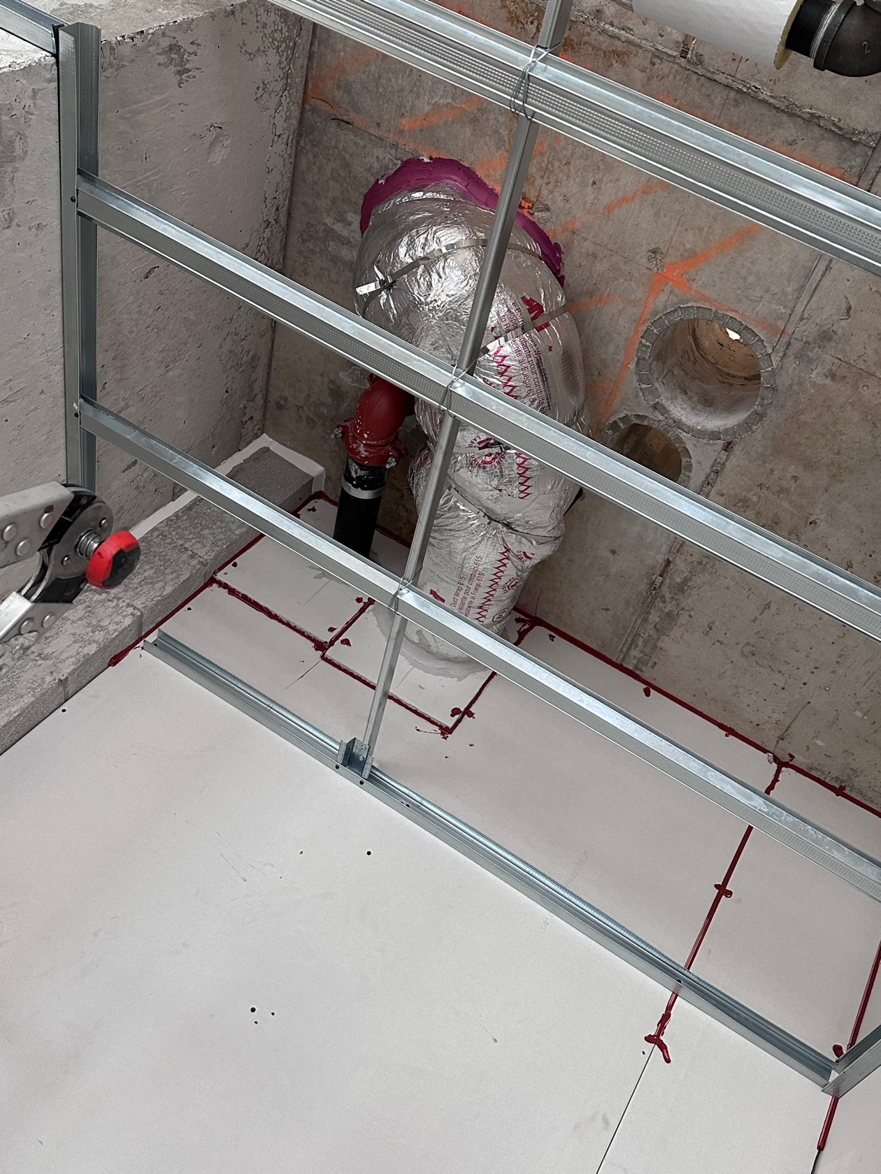710114a223d8dad0d2b393ca84a171fd4be79ef6acdec4115b4215eb574bed2e Fire-rated pipe wrap on a vertical riser with red fire barrier sealant passing through a floor slab, seen through metal stud framing, installed by Metro Insulation in a Toronto high-rise