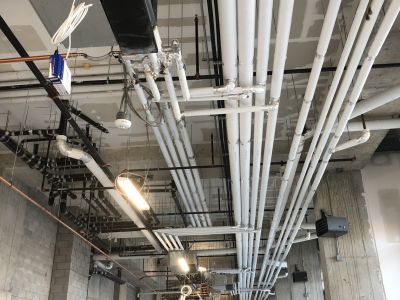 Exposed HVAC fan coil unit with copper coils and insulated pipes installed by Metro Insulation Exposed HVAC fan coil unit with copper coils beside white PVC-jacketed insulated pipes and a black fire pipe in a ceiling space
