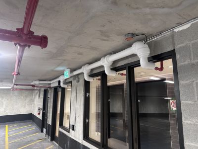 White PVC-jacketed insulated pipe runs across a concrete ceiling installed by Metro Insulation White PVC-jacketed insulated pipes running across a concrete ceiling beside a red fire pipe and cinder block wall