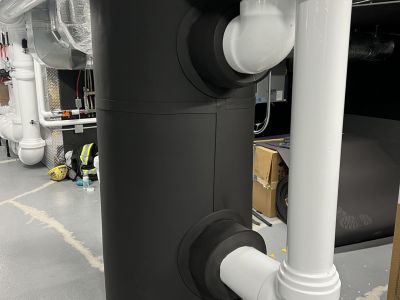 Large white PVC-jacketed insulated pipe elbows around an expansion tank installed by Metro Insulation Large white PVC-jacketed insulated pipe elbows and fittings around a black expansion tank with foil-faced insulation behind